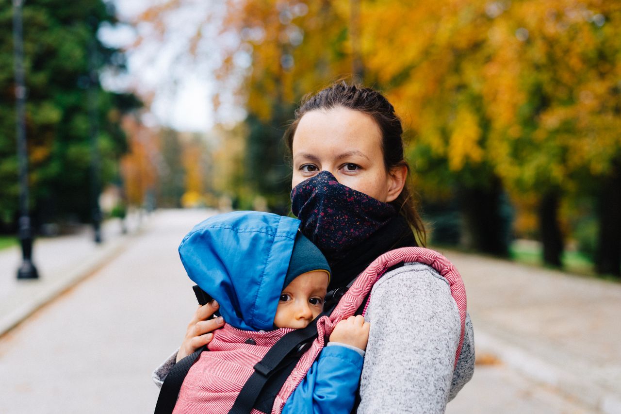 team-03 Woman wearing a mask carrying a baby in a carrier during a fall walk in the park.