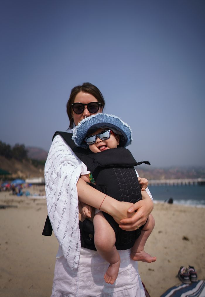 services-01 Young mother with adorable smiling baby in sunglasses and hat in baby carrier on sandy beach in sunny weather looking at camera