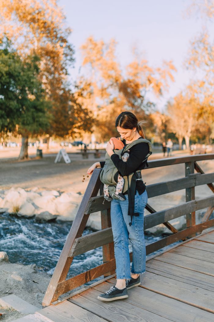services-02 Woman holding baby in a carrier while standing on a wooden bridge in a park during autumn.